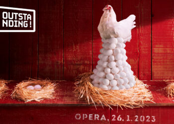 A white hen sits atop a huge nest of eggs in a red hen house.  The surrounding nest have only one or two eggs.