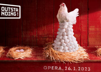 A white hen sits atop a huge nest of eggs in a red hen house.  The surrounding nest have only one or two eggs.