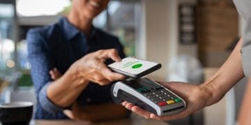 Woman paying bill through smartphone using NFC technology in a restaurant. Satisfied customer paying through mobile phone using contactless technology. Closeup hands of mobile payment at a coffee shop